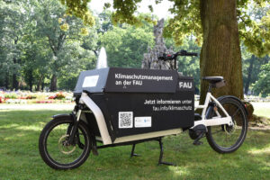 Ein Foto von einem Lastenrad mit schwarzem Transportkasten im Schlossgarten mit einem Baumstamm und dem Springbrunnen im Hintergrund.